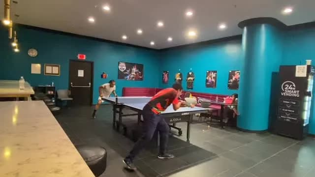 Framed table tennis posters line the teal wall above the bench seating, while two players rally at the near table