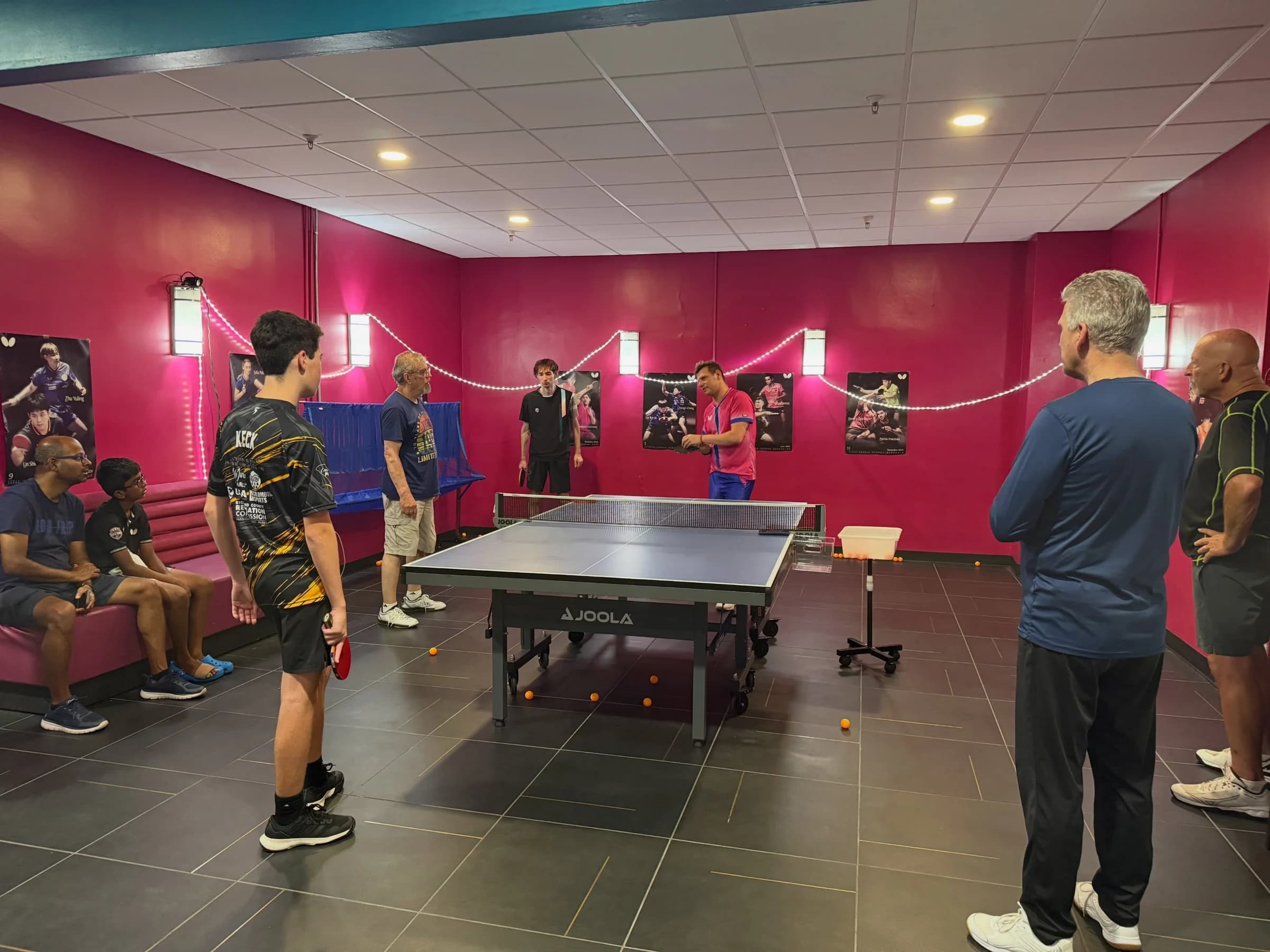 Five players of different ages stand at a blue table in the teal-walled room, paddles up